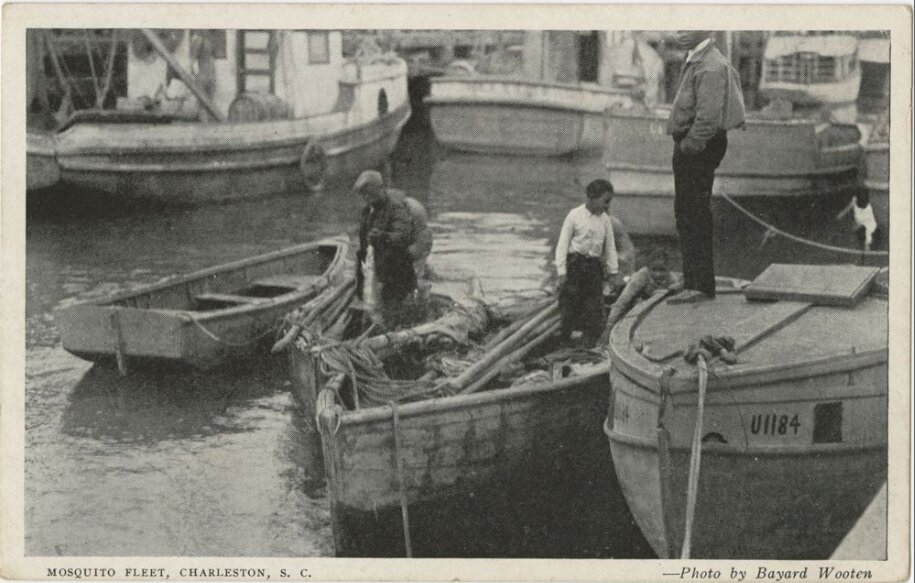 Early twentieth-century postcard photograph of men working around small open fishing boats moored in Charleston harbor, with larger boats behind.