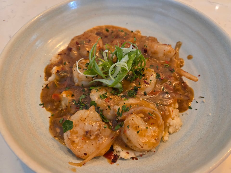 Sautéed shrimp over smoked gouda grits and tasso gravy, topped with shredded scallion greens and pepper flakes in a pale blue bowl.