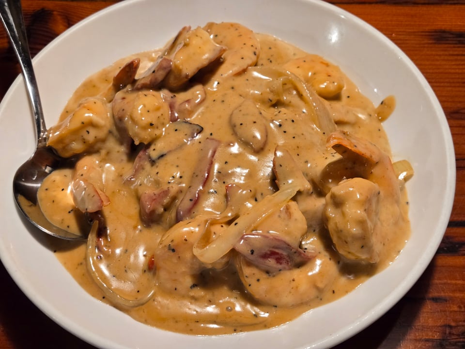 Close view of shrimp with tails, onions, and red pepper pieces covered in orange sherry cream sauce on a white plate.