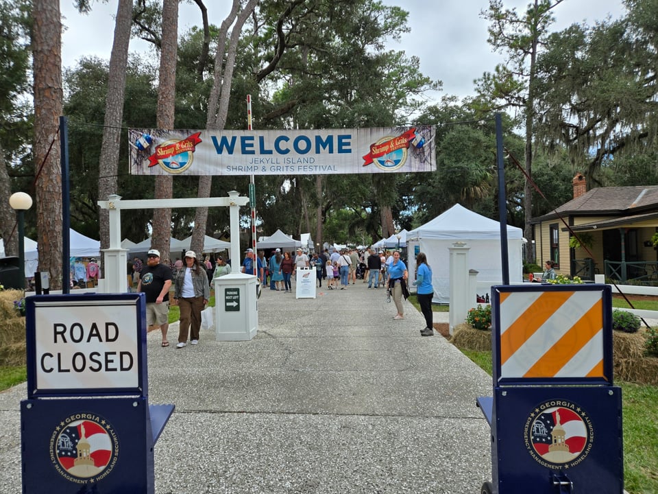 A pedestrian entrance lined with festival tents under live oaks and pines, with a banner reading Welcome, Jekyll Island Shrimp & Grits Festival, strung over the path between two road-closed barricades.