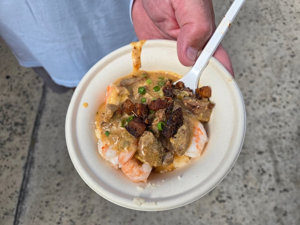 A hand holding a bowl of shrimp over grits under a brown gravy with sausage, crisp pork pieces, and scallion, with a white plastic fork resting in the bowl.