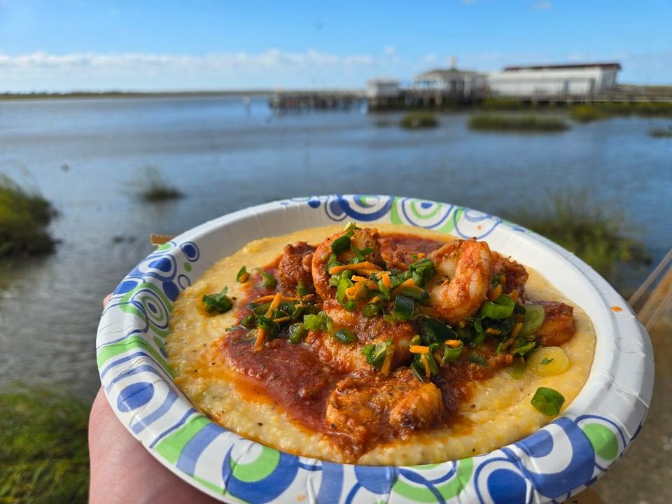 A hand holding a paper bowl of shrimp and grits in a red tomato gravy, topped with chopped scallion and shredded carrot, in front of a tidal creek and a pier.