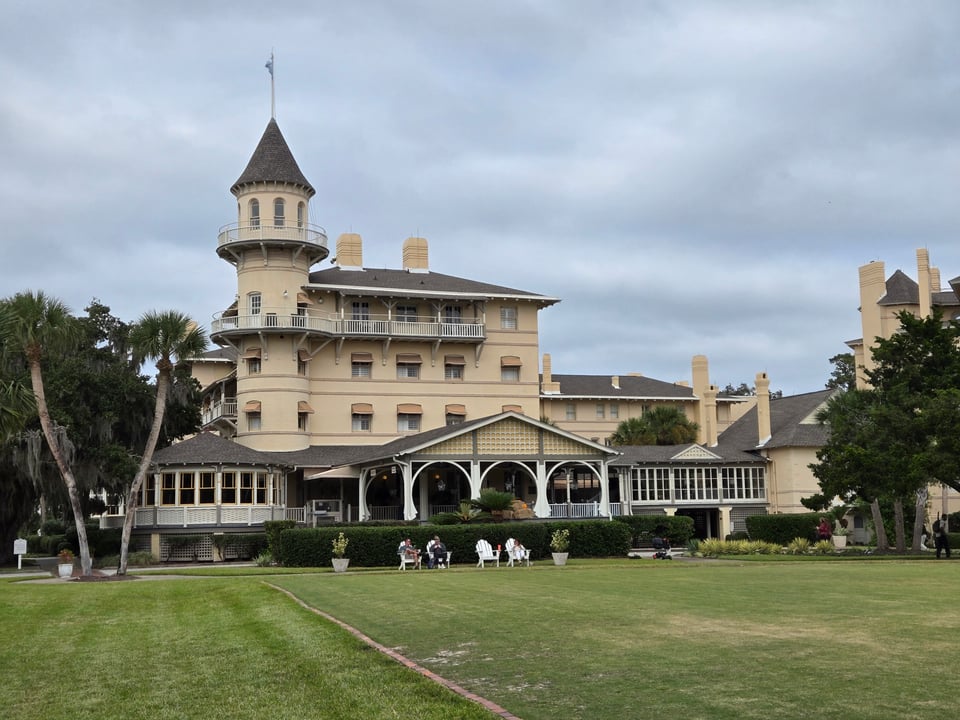 The turreted, pale-yellow Jekyll Island Club hotel with a wraparound porch behind a broad green lawn under a cloudy sky.
