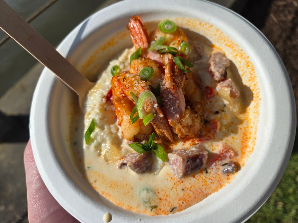 A compostable bowl of creamy grits topped with shrimp with the tails still on, sausage pieces, and scallion in a pale orange sauce, held with a wooden fork in the bowl.