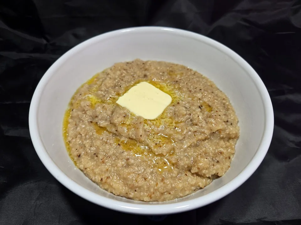 A white bowl of dark-toasted grits with a pat of butter melting on top and golden butter pooling around the edge