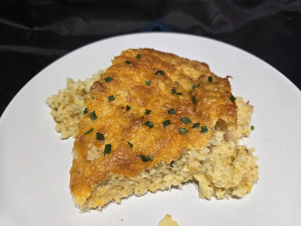 A serving of baked cheesy grits on a white plate, with a golden cheese crust and chives over soft, fluffy grits