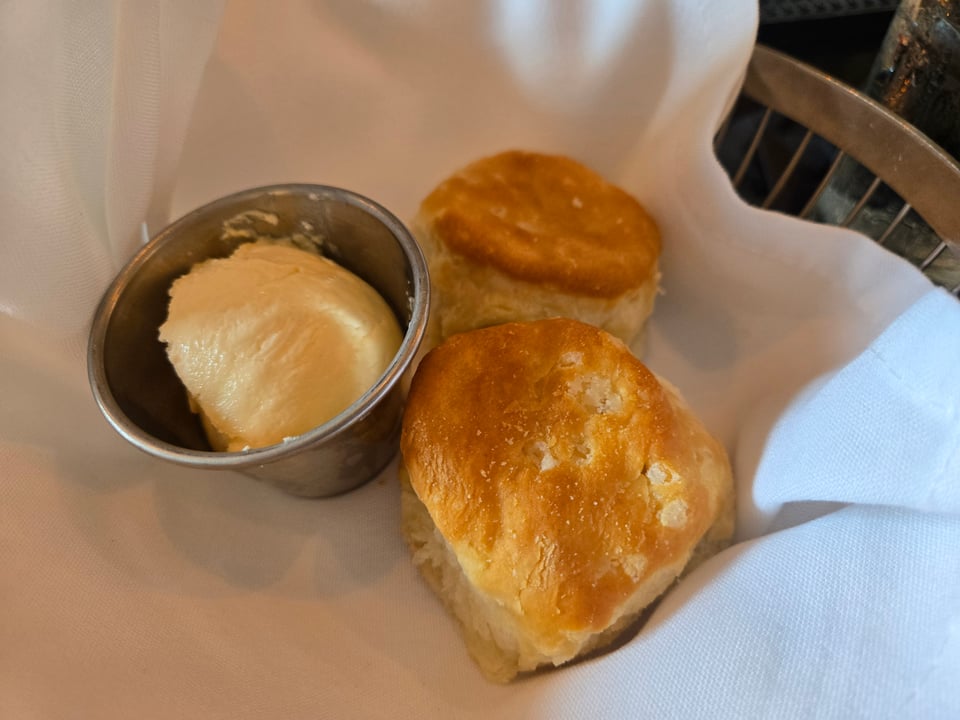 Two golden biscuits beside a metal cup of butter in a cloth-lined basket.